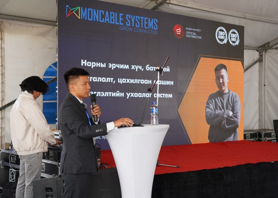 Костюм өмссөн эр тайзан дээр микрофон барин үг хэлж байна. Түүний ард “Moncable Systems” компанийн лого нь ухаалаг барилгын тухай монгол бичээс болон гараа зөрүүлэн зогсож буй өөр нэг хүний гэрэл зургийн хамт байрлуулсан байна. - Business.MN