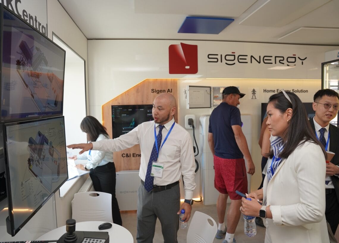 Цагаан цамцтай, зангиатай эр хоёр том дэлгэц рүү чиглэн, технологийн үзэсгэлэн дээр бизнесийн хувцастай хоёр хүнд ногоон ирээдүйн шийдлийг тайлбарлаж байна. Бусад оролцогчид болон ухаалаг барилга дэлгэцийн ард харагдаж байна. - Business.MN