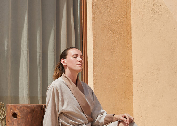 Хөнгөн дээлтэй эмэгтэй орчин үеийн шаргал барилгын гадаа бетонон хашаан дээр тухлан сууж, нүдээ аниад наранд жаргах нь тогтвортой аялал жуулчлалыг тусгах тайван агшин. Түүний хажууд нэхмэл малгай, модон сандал; цонхигор хөшиг шилэн хаалгыг бүрхэнэ. - Business.MN