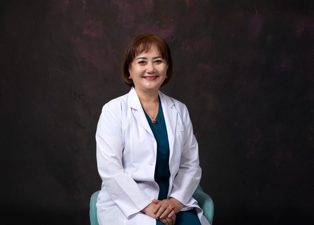 Шүдний эмнэлэгийнх бололтой цагаан лабораторийн цув, цайвар өнгийн скраб өмссөн богино бор үстэй эмэгтэй бараан өнгийн алаг дэвсгэр дээр цайвар хөх өнгийн сандал дээр инээмсэглэн сууж байна. - Business.MN