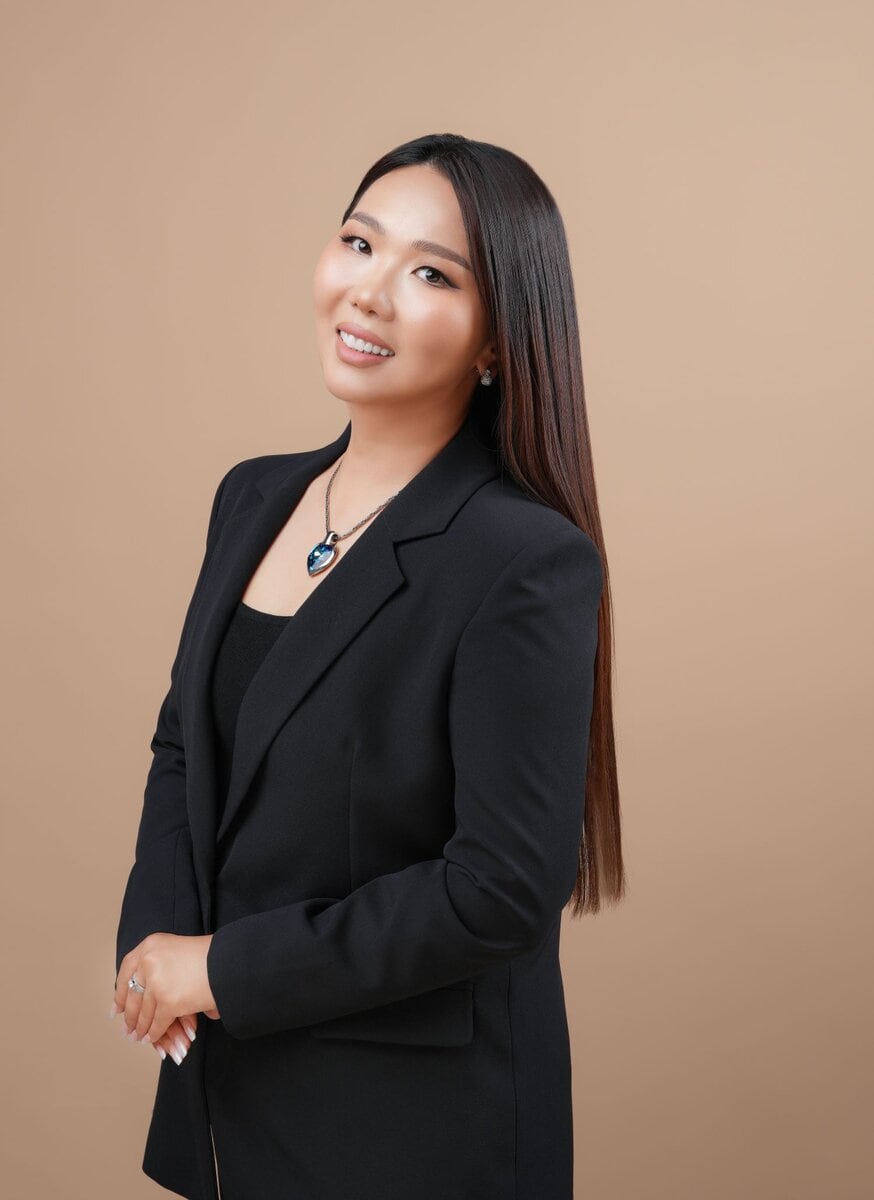 Хар пиджак өмсөж, хүзүүний зүүлт зүүсэн урт шулуун үстэй эмэгтэй энгийн шаргал дэвсгэр дээр инээмсэглэн зогсож, гараа урдуур нь зөөлөн атгаж, тэнцвэртэй амьдралыг илтгэнэ. - Business.MN