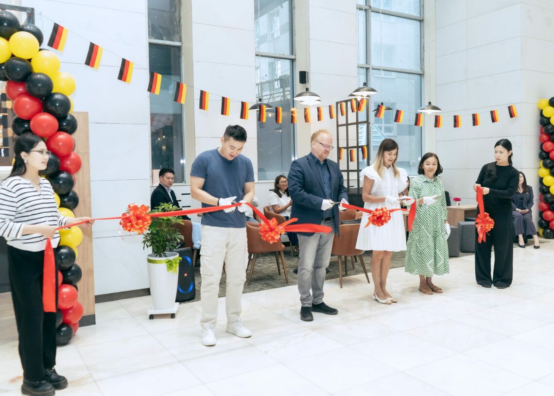 Хар, улаан, шар өнгийн бөмбөлөг, туг далбаагаар чимэглэсэн танхимын арга хэмжээнд зургаан хүн дараалан зогсоод улаан тууз хайчилж байна. Хэд хэдэн үзэгчид ард сууж байна. - Business.MN