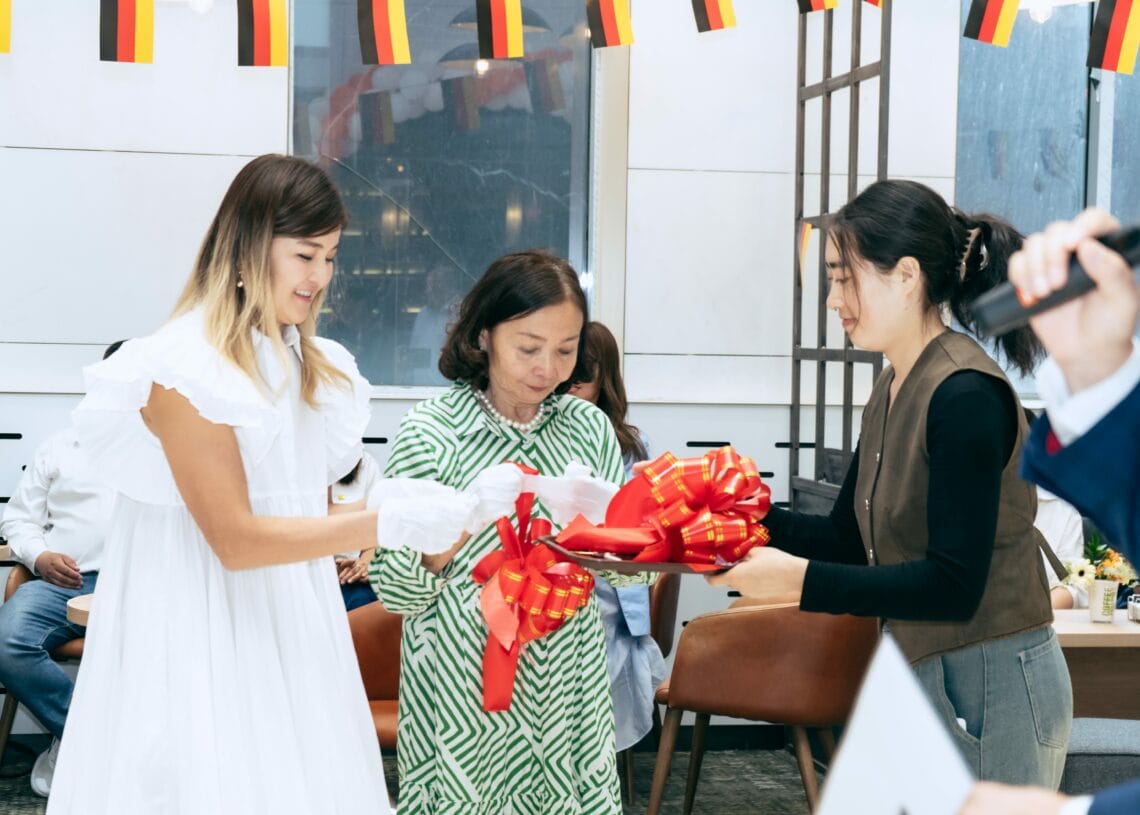 Гурван эмэгтэй улаан, шар өнгийн туузан нум барин доторх тууз хайчлах ёслолд оролцож байна. Тэдний дээр Германы далбаа өлгөөтэй, ард нь хүмүүс сууж байна. - Business.MN