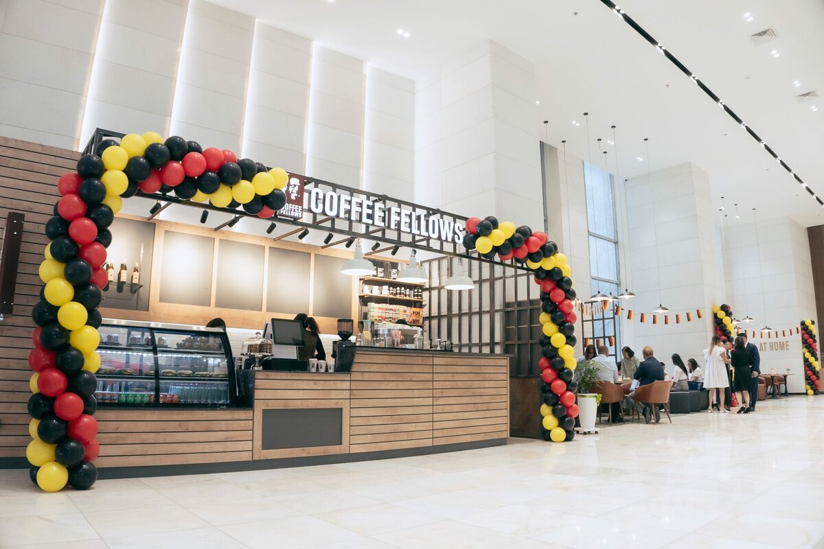 Улаан, шар, хар өнгийн бөмбөлөг нуман хаалгаар чимэглэсэн орчин үеийн кофены мухлагыг хүмүүс ширээний ард сууж, ярилцах цэлгэр, гэрэлтэй үүдний танхимд байрлуулсан. - Business.MN