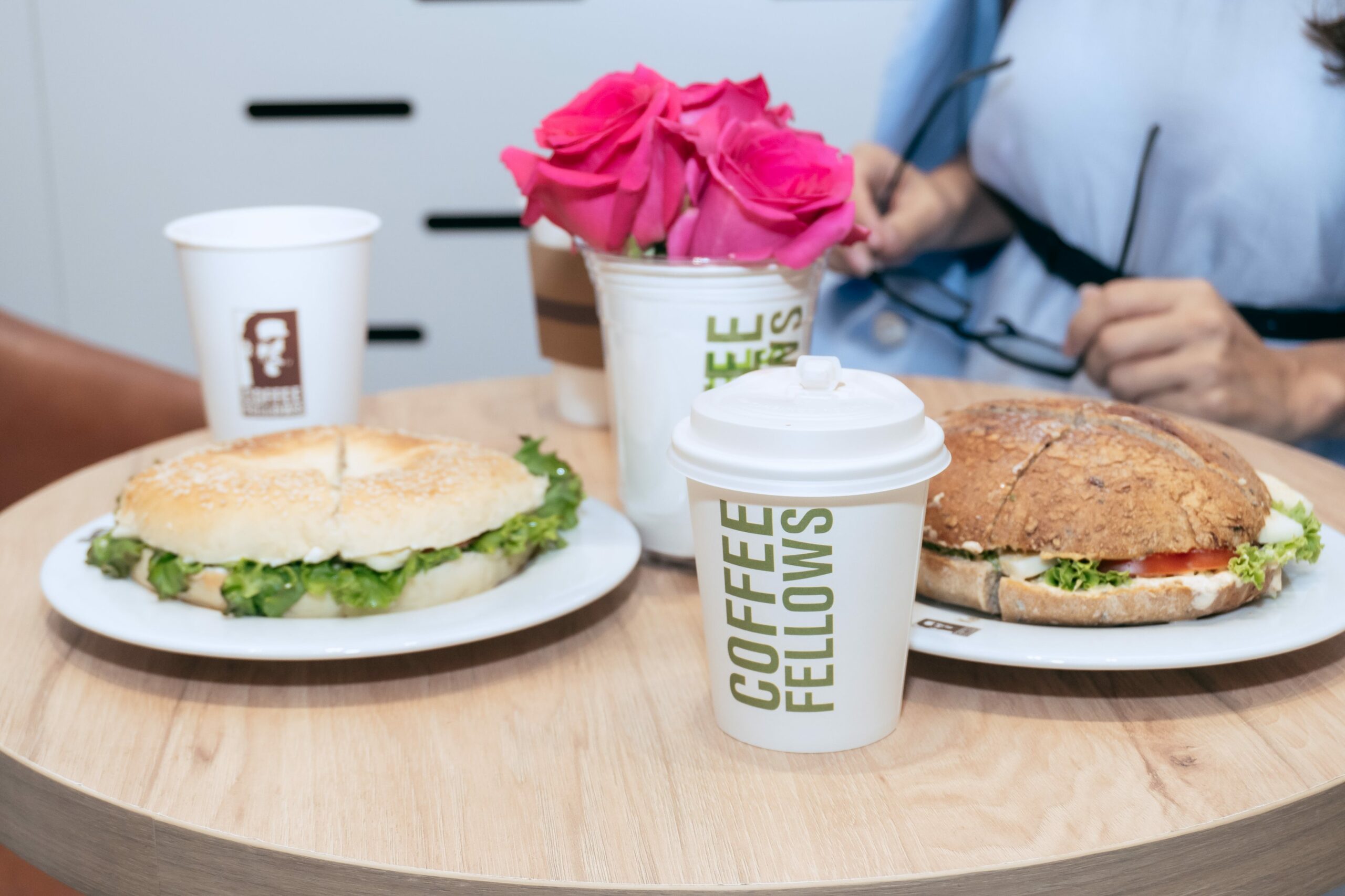 Тавган дээрх хоёр хачиртай талх, "Кофены хамт олон" гэсэн шошготой хоёр кофены аяга, ягаан сарнайн баглаа бүхий дугуй ширээ. Цаана нь нүдний шил барьсан хүн хэсэгчлэн харагдана. - Business.MN