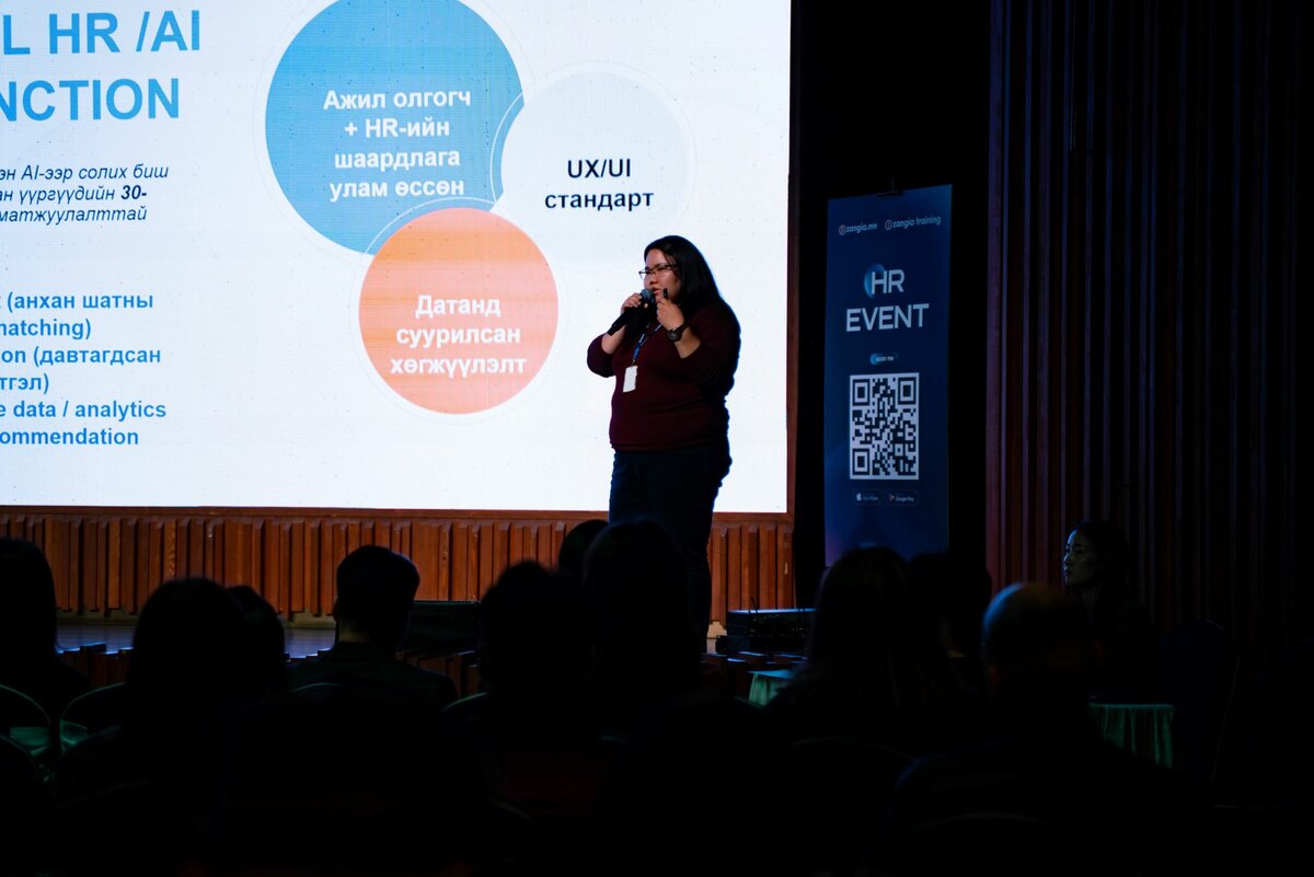 Тайзан дээр зогсож буй эмэгтэй HR EVENT дээр микрофоноор ярьж байгаа бөгөөд ард нь текст, диаграммуудыг харуулсан дэлгэц, урд нь үзэгчид хоорондоо сүлжээгээ үүсгэсэн байна. - Business.MN