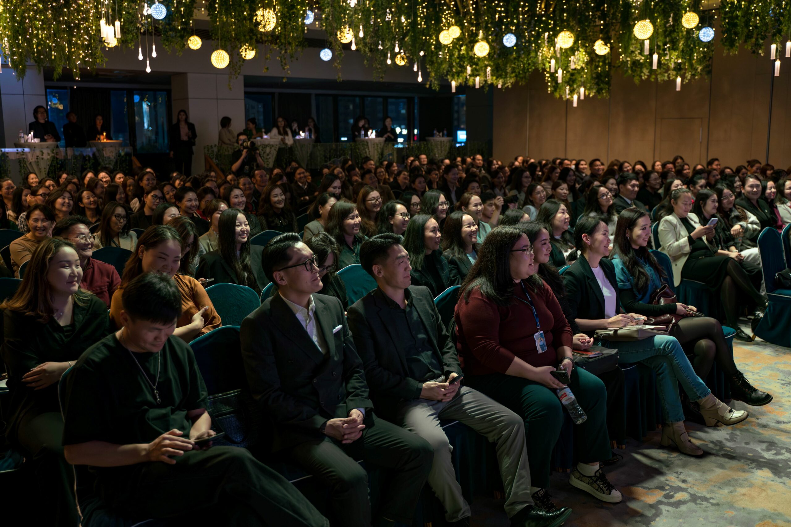 Өргөгдсөн гэрэл, ногоон байгууламж бүхий тохижуулсан танхимд олон үзэгчид анхааралтай сууж байна. Ихэнх хүмүүс, магадгүй хүний нөөцийн мэргэжилтнүүд урагшаа харж, үл үзэгдэх үйл явдал эсвэл илтгэгчийг харж байна. - Business.MN