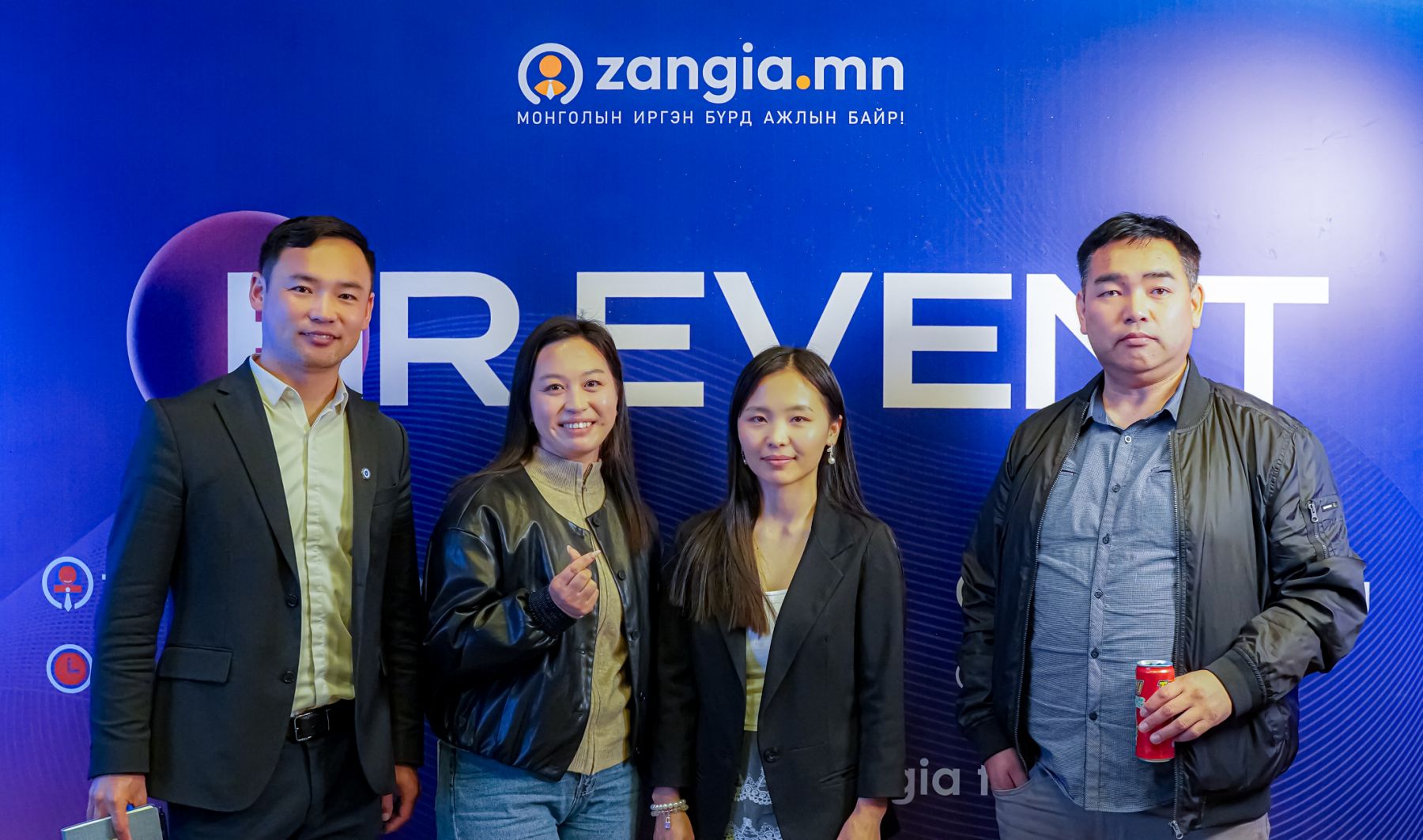 Цэнхэр “zangia.mn” EVENT фонны өмнө гурван эмэгтэй, нэг эрэгтэй дөрвөн хүн инээмсэглэн зогсож байна. Нэг нь ундааны лааз барьдаг; others have hands in pocket or gestu, highlighting хүний нөөцийн салбарын уур амьсгалыг. - Business.MN