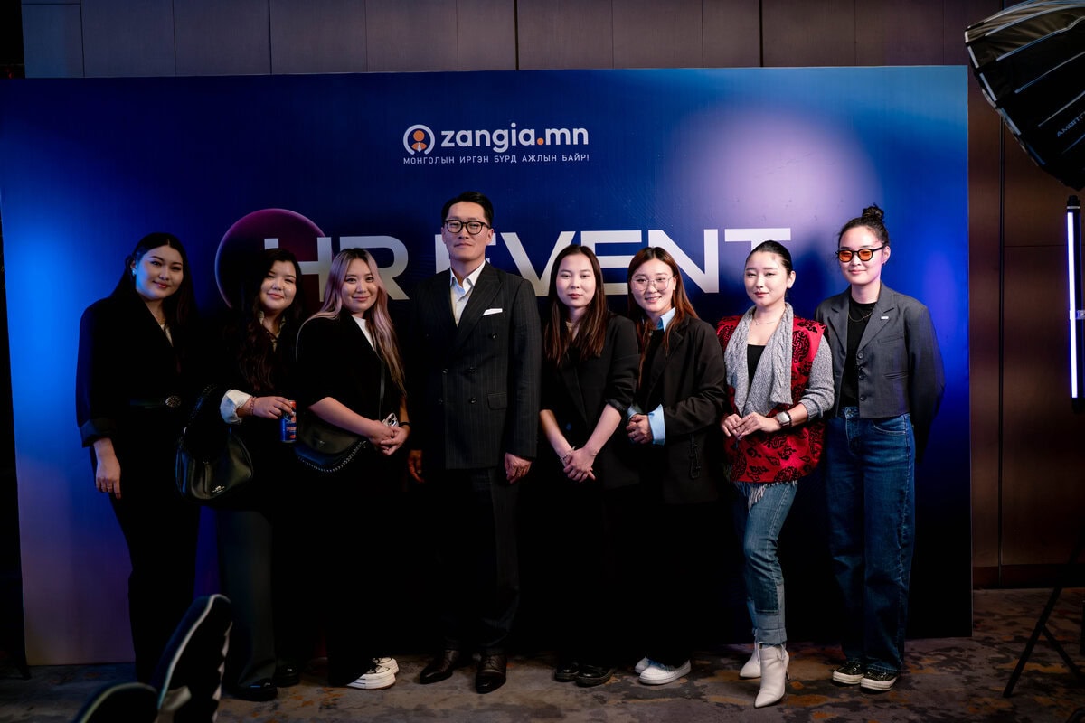 Цэнхэр, нил ягаан өнгийн "zangia.mn хүний нөөц HR EVENT"-ийн тайзны өмнө ажил хэрэгч, энгийн хувцас өмссөн найман хүн хамтдаа зогсож гэрэл зураг авахуулахаар инээмсэглэж байна. - Business.MN
