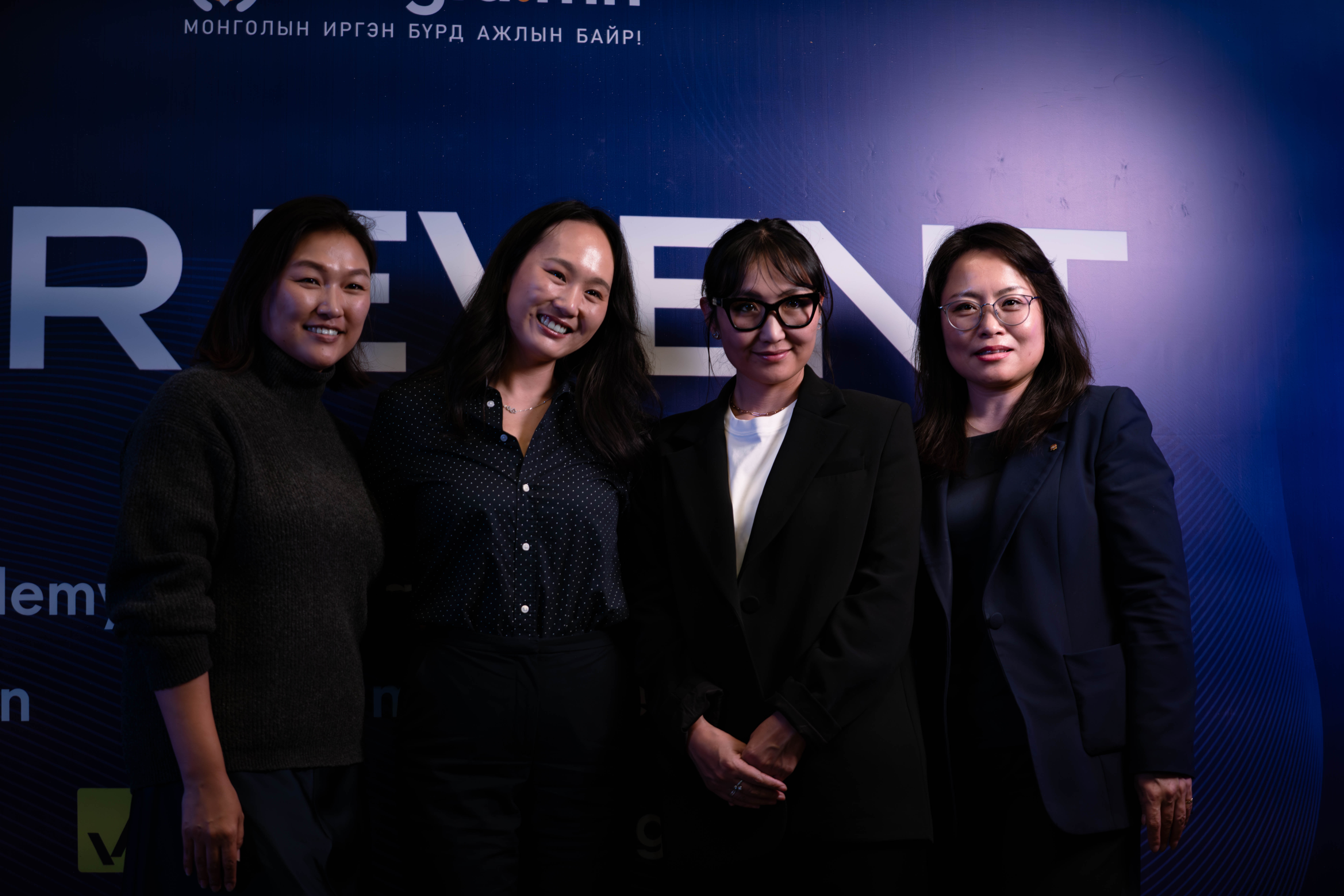 Ажил хэрэгч эсвэл ухаалаг энгийн хувцас өмссөн дөрвөн эмэгтэй хүний нөөцийн арга хэмжээ эсвэл мэргэжлийн цугларалтад оролцож буй агшин зуурын тексттэй хар хөх өнгийн дэвсгэр дээр инээмсэглэн гэрэл зураг дарж байна. - Business.MN