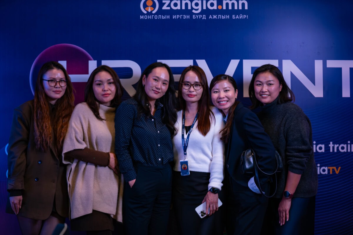 Зургаан эмэгтэй хамт зогсож, "EVENT" гэсэн хэсэгчлэн харагдахуйц монгол бичвэр бүхий цэнхэр өнгийн арын дэвсгэр дээр хамтдаа зургаа авахуулж байна. Тэд ажил хэрэгч эсвэл ухаалаг энгийн хувцас өмссөн нь мэргэжлийн арга хэмжээнд хүний нөөцийн ач холбогдлыг онцолж байна. - Business.MN