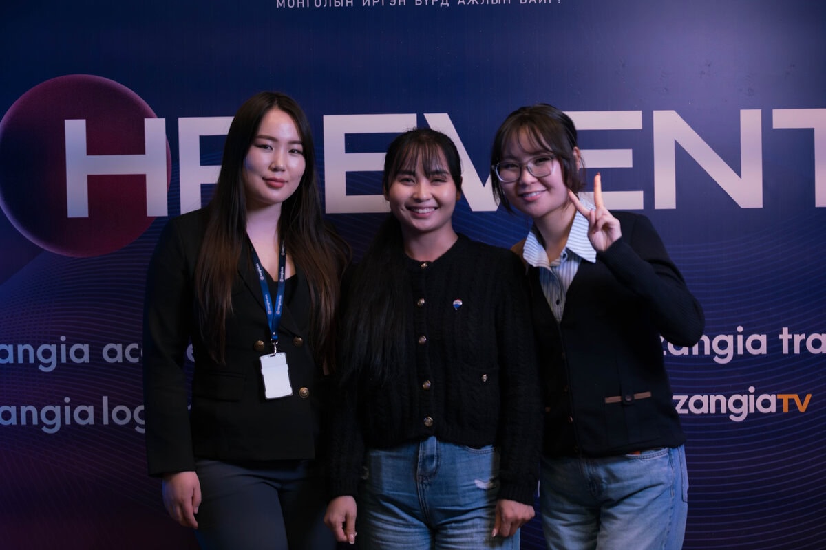 Гурван залуу эмэгтэй том цагаан бичвэр, компанийн лого, хүний нөөцийн брэнд бүхий хар хөх өнгийн арга хэмжээний арын дэвсгэрийн өмнө инээмсэглэн зогсож байна. Баруун талд байгаа нэг эмэгтэй гараараа энх тайвны тэмдэг хийж байна. - Business.MN