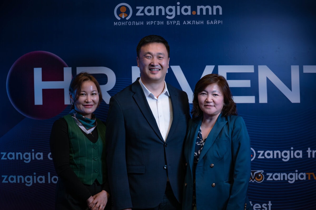 Хүний нөөцийг онцолж "zangia.mn", "HR Event" гэсэн бичээс бүхий цэнхэр дэвсгэрийн өмнө гурван хүн инээмсэглэн зогсож байна. Хоёр эмэгтэй хар хувцастай, ажил хэргийн хувцас өмссөн эрэгтэйн хоёр талд зогсож байна. - Business.MN