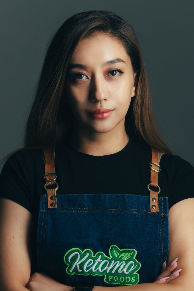 Урт бор үстэй эмэгтэй ногоон өнгөөр хатгамал "Кетомо Фүүдс" бүхий хар цамц, жинсэн хормогч өмссөн байна. Тэрээр саарал дэвсгэрийн эсрэг итгэлтэйгээр зогсч, тухлаг кафе болон Ketomo Cafe Wine-ийн зочломтгой, гэртээ мэт мэдрэмжийг илэрхийлдэг. - Business.MN