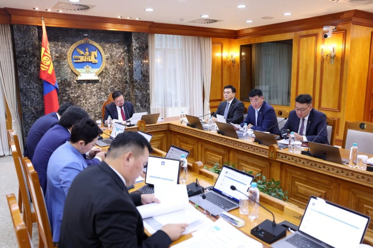 Төрийн албан хаагчид хувцастай, том модон хурлын ширээний ард зөөврийн компьютер, бичиг баримттай сууж, хананд нь Монгол Улсын төрийн сүлдтэй төрийн албан ёсны танхимд 2026 оны төсвийн талаар хэлэлцэж байна. - Business.MN