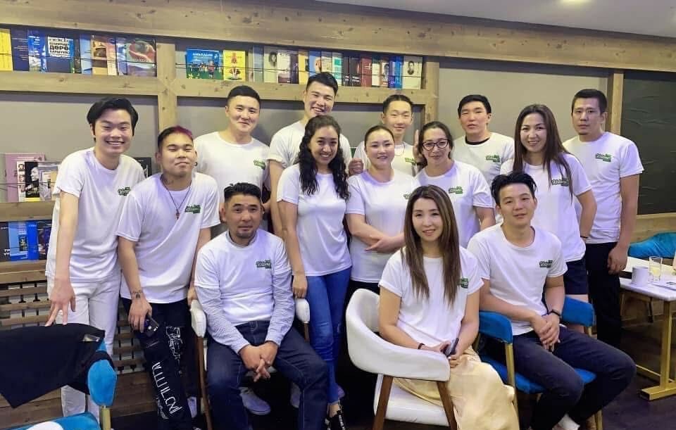 Арван гурван хүний бүрэлдэхүүнтэй, цагаан цамц өмссөн хүмүүс номын тавиур, модон чимэглэл бүхий Ketomo Cafe Wine-ийн тухтай өрөөнд хамтдаа зургаа авахуулж, гэртээ байгаа мэт сэтгэгдэл төрүүлж байна. Ихэнх нь зогсож байхад цөөхөн хэд нь камерын өмнө инээмсэглэн сууж байна. - Business.MN