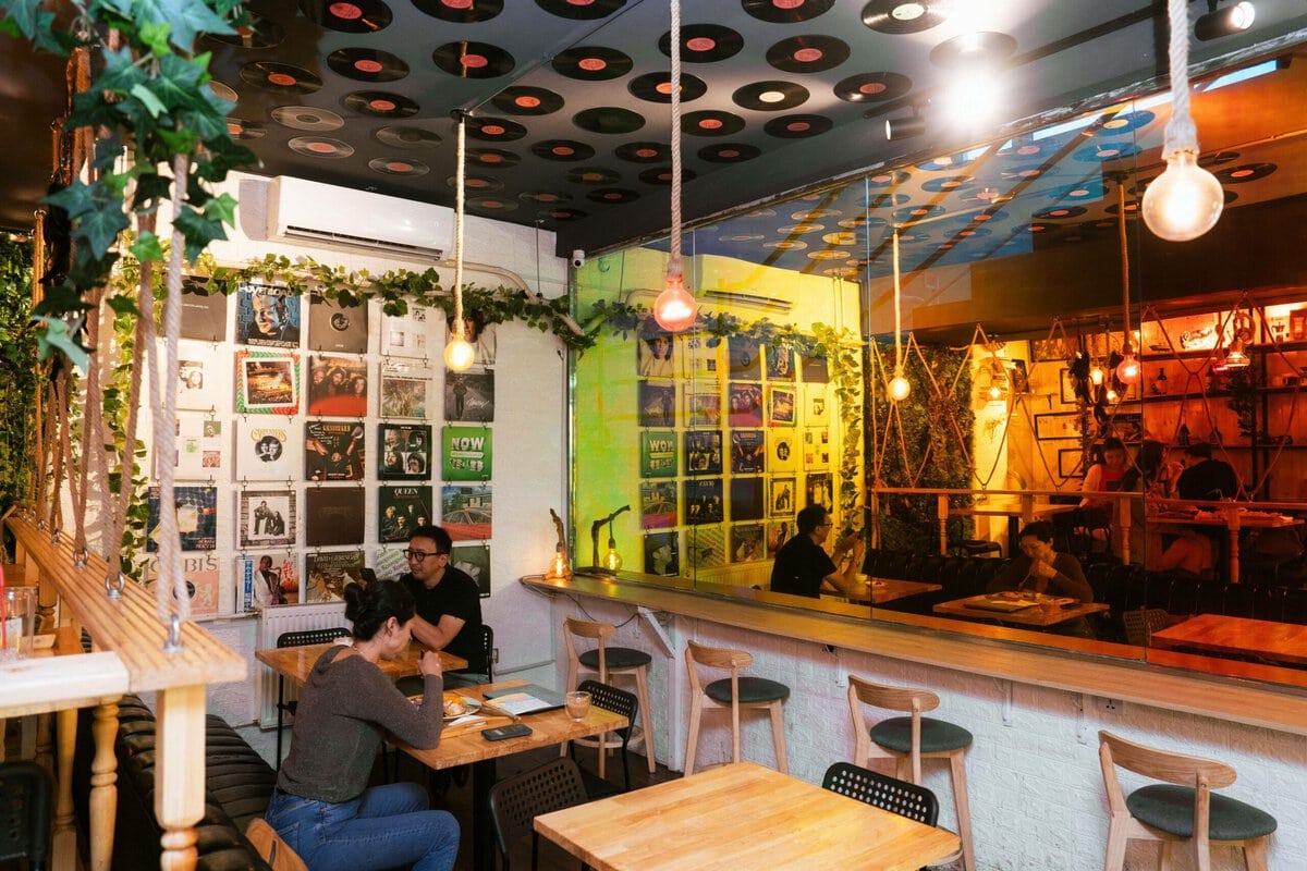 Модон ширээ, таазанд винил пянз, ханан дээр хөгжмийн цомгийн хавтастай тухтай тухлаг кафе. Хоёр хүн ширээний ард дарс шимэн сууж байхад бусад нь бааранд сууж байна. Дулаан гэрэлтүүлэг, өлгөөтэй ургамал нь тайван уур амьсгалыг бүрдүүлдэг. - Business.MN