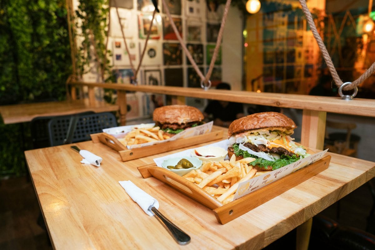 Ширээн дээрх хоёр модон тавиур, дээр нь хайлсан бяслаг, шанцайны ургамал, улаан лооль бүхий бургер, шарсан төмс, даршилсан өргөст хэмхтэй хамт үйлчилнэ. Ногоон байгууламж, дулаан гэрэлтүүлгээр дүүрсэн тухтай Ketomo Cafe Wine-д гэртээ ийм туршлагыг мэдрээрэй. - Business.MN