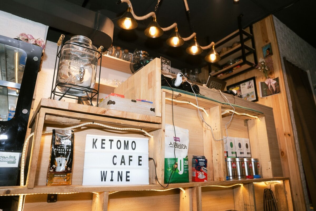 Кофе, дарсны бүтээгдэхүүн дэлгэн тавьсан модон тавиур, ундааны шилэн сав, өлгөөтэй гэрэл, "KETOMO CAFE WINE" гэсэн бичигтэй гэрлийн хайрцаг бүхий тохилог, тухлаг кафены интерьер нь Ketomo Cafe Wine-г гэртээ үзэхэд тохиромжтой. - Business.MN