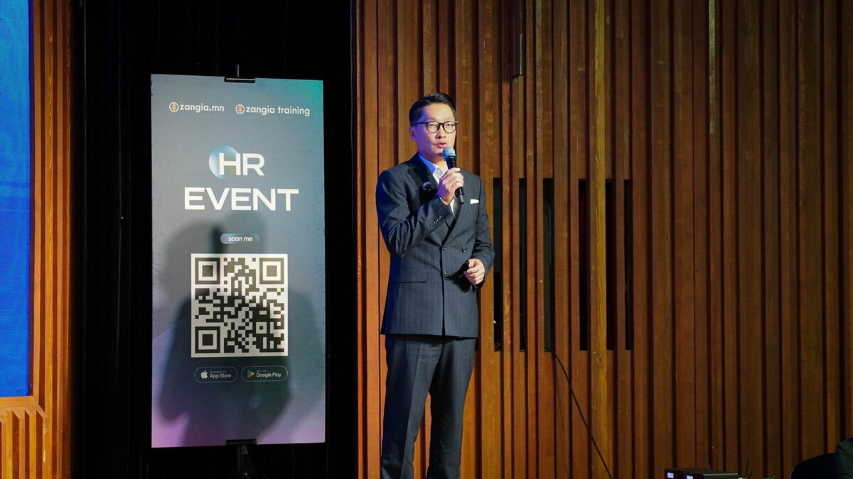 Костюм өмссөн хүн HR EVENT-ийн тайзан дээр QR код бүхий самбар, хүний нөөцийн сүлжээнд зориулсан олон нийтийн мэдээллийн хэрэгслийн хажууд микрофоноор ярьж байна. Арын дэвсгэр нь босоо модон хавтангуудыг харуулж байна. - Business.MN
