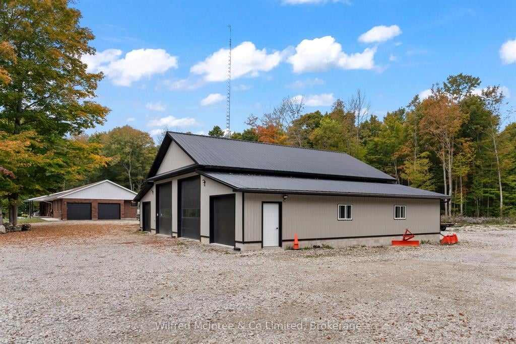 A modern metal garage building with black trim sits on a gravel lot, surrounded by trees. A smaller building and a three-car garage are visible in the background.