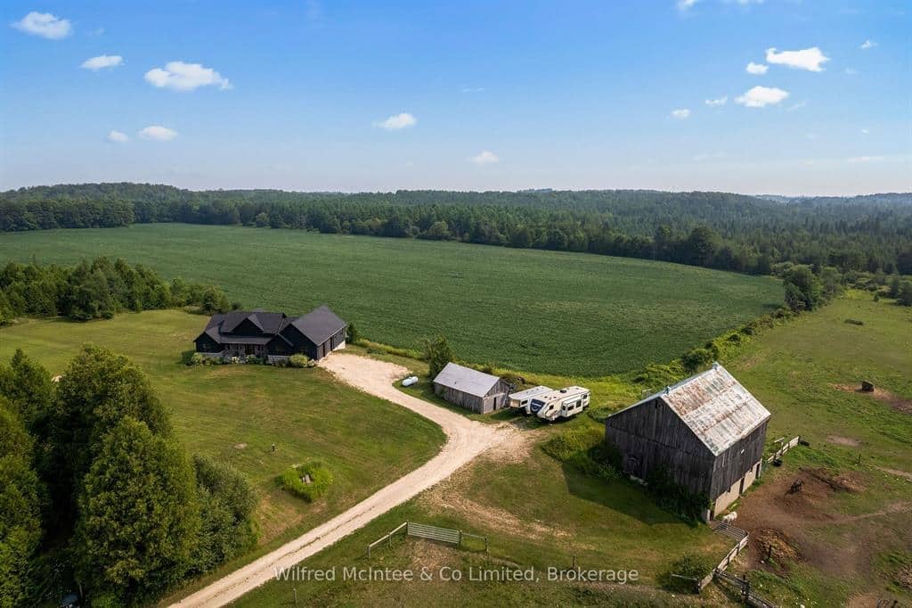 Aerial view of a rural property with a house, barn, shed, RV, and large green fields surrounded by trees under a blue sky.