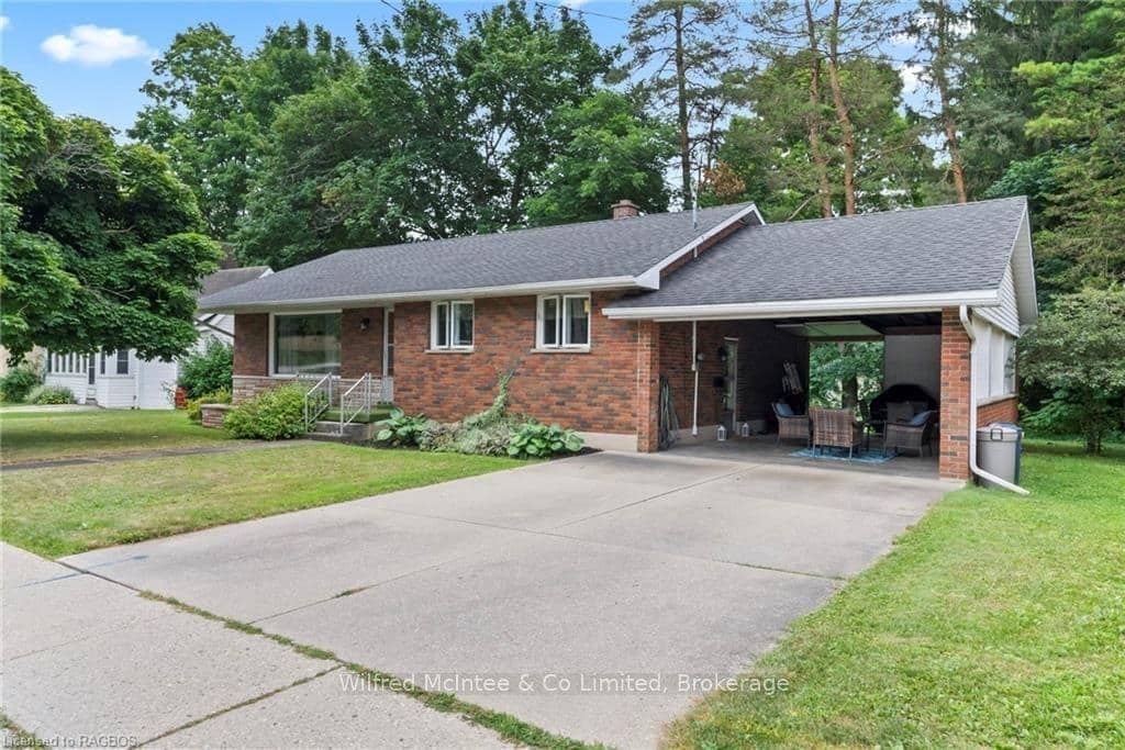 Single-story brick house with a covered carport, paved driveway, front lawn, and mature trees in the background.