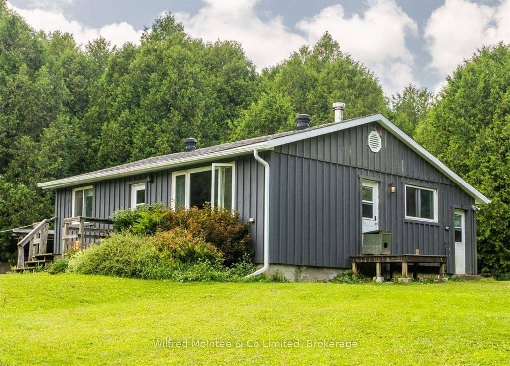 Single-story gray house with a sloped roof, surrounded by grass and shrubs, set against a background of dense green trees under a cloudy sky.