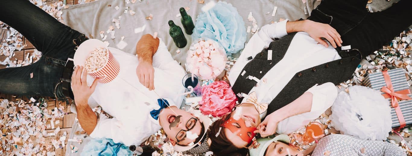 Vier Menschen liegen auf dem Boden, umgeben von Konfetti und festlicher Dekoration. Sie tragen Fliegen und Masken. Es ist eine Szene wie bei einer lebhaften Firmenfeier. Zwei halten Popcorn und Snacks in den Händen, umgeben von Geschenkkartons, die dem Fest noch mehr Charme verleihen.