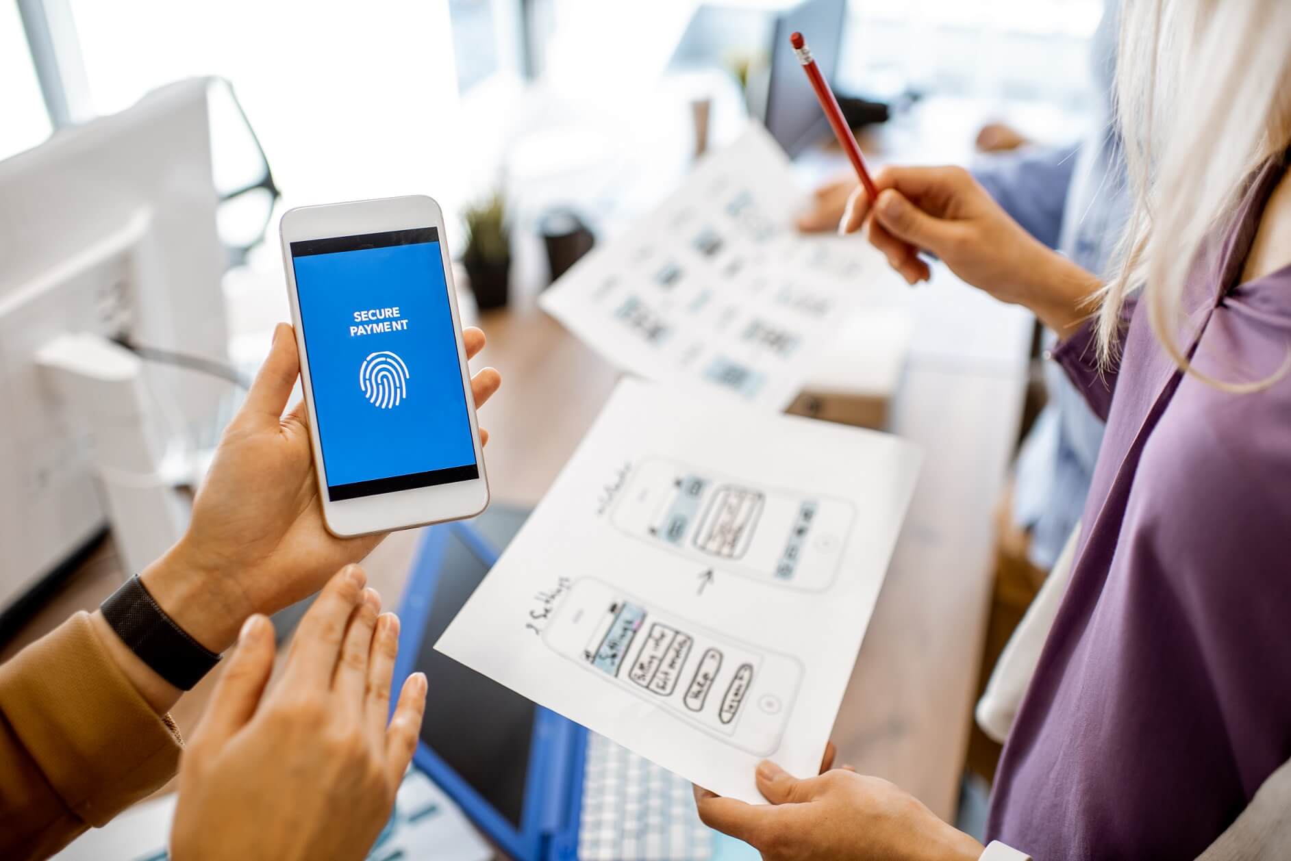 A person holding a smartphone displaying a "Secure Payment" screen and fingerprint icon. Another person points to a paper showing DigiComet's mobile app designs during a discussion in a modern office setting.