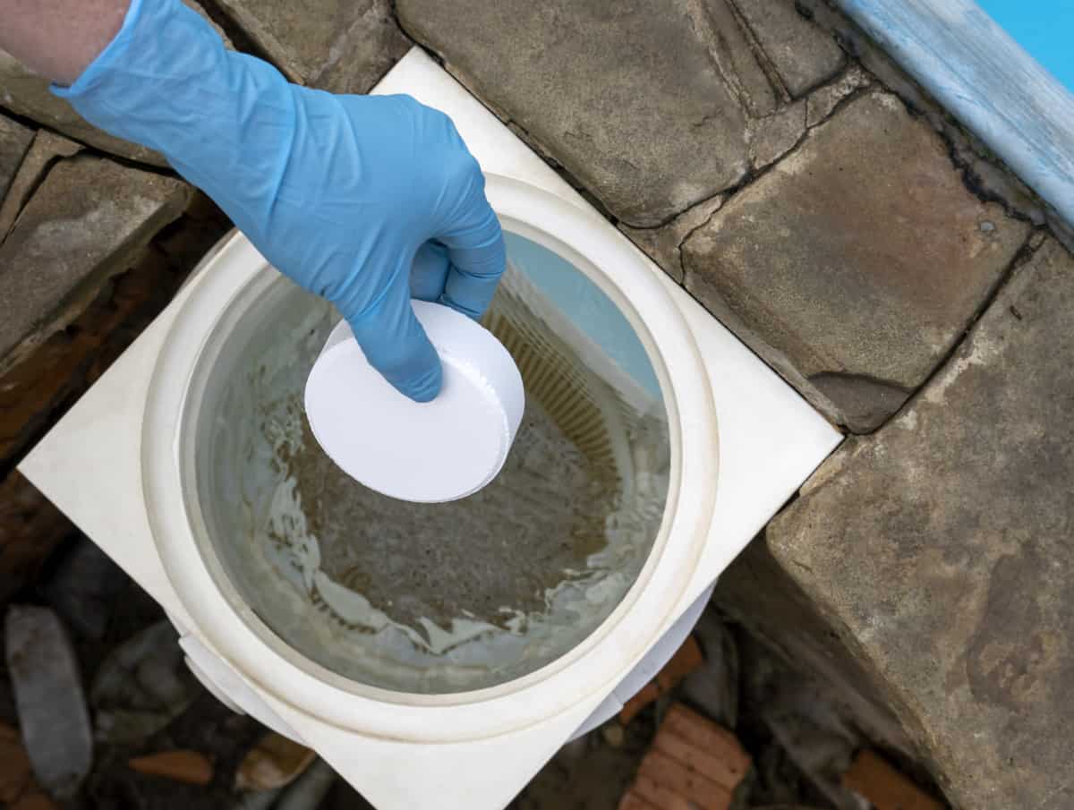 A hand with blue gloves holding a 3 inch chlorine tablet in a skimmer.