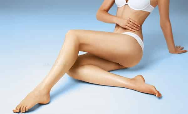 O femeie în lenjerie intimă albă stă pe podea cu picioarele întinse, cu o mână pe stomac, pe un fundal alb-albastru în gradient, evocând atmosfera senină a unei sesiuni Remodelare Corporala Iași.