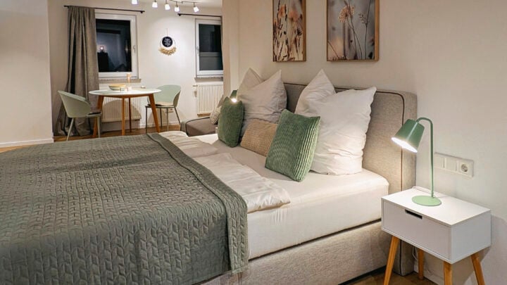 MainZauber Apartments früher MainTerrasse Miltenberg am Main - Entdecken Sie die perfekte Ferienwohnung für Ihren nächsten Kurzurlaub. Diese luxuriöse Unterkunft verfügt über ein gemütliches Schlafzimmer mit einem ordentlich gemachten Bett, das mit eleganter grün-weißer Bettwäsche ausgestattet ist. Genießen Sie den Komfort von zwei Nachttischen mit Lampen, die für einen angenehmen Aufenthalt sorgen. Genießen Sie Ihre Mahlzeiten am kleinen Esstisch mit Stühlen in der Nähe des Fensters, das Ihnen beim Essen eine einladende Aussicht bietet. Die neutrale Einrichtung der Wohnung, ergänzt durch geschmackvolle Wandkunst und sanfte Beleuchtung, schafft eine warme und einladende Atmosphäre, die ideal zum Entspannen während Ihres Urlaubs ist.