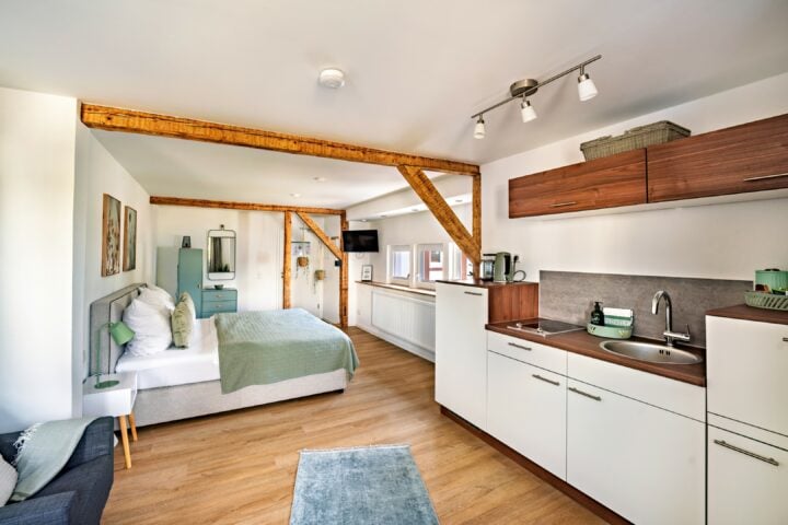 MainZauber Apartments Miltenberg am Main - Entdecken Sie Ihre perfekte Ferienwohnung mit unserem bezaubernden modernen Studio, ideal für alle, die eine luxuriöse Unterkunft suchen. Diese gehobene Ferienwohnung bietet ein gemütliches Bett unter eleganten Holzbalken und eine voll ausgestattete Küchenzeile, die perfekt für Ihren Aufenthalt ist. Der Innenraum empfängt Sie mit stilvollen Weiß- und Holztönen, ergänzt durch einen schicken Teppich auf dem polierten Holzboden und exquisite Wandkunst. Genießen Sie das natürliche Licht, das durch das Fenster strömt, und genießen Sie Unterhaltungsmöglichkeiten mit einem eleganten, an der Wand montierten Fernseher. Erleben Sie luxuriöses Wohnen in dieser anspruchsvollen Ferienwohnung!