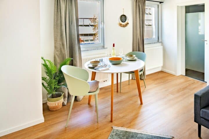 MainZauber Apartments Miltenberg am Main - Entdecken Sie den perfekten Kurzurlaub in unserer luxuriösen Ferienwohnung, wo Komfort auf Stil trifft. Genießen Sie Ihre Mahlzeiten oder entspannen Sie bei einem Glas Wein in einer charmanten Essecke für zwei Personen. Diese gemütliche Unterkunft verfügt über einen runden Tisch mit elegantem Geschirr und zwei stilvollen hellgrünen Stühlen. Der einladende Raum verfügt über schöne Holzböden und wird durch weiche, graue Vorhänge an den Fenstern aufgewertet, die eine ruhige Atmosphäre schaffen. Eine kleine Topfpflanze verleiht einen Hauch von Natur, während ein geschmackvoller Wandkranz dieser exquisiten Umgebung Charakter verleiht. Gönnen Sie sich das ultimative Urlaubserlebnis in diesem wunderschön eingerichteten Rückzugsort, wo jedes Detail darauf ausgelegt ist, Ihren Aufenthalt unvergesslich zu machen.