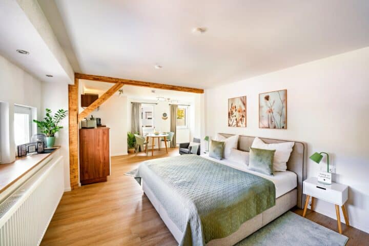 MainZauber Apartments Miltenberg am Main - Entdecken Sie die perfekte Ferienwohnung für Ihren Kurzurlaub. Sie verfügt über ein luxuriöses Schlafzimmer mit eleganter grün-weißer Bettwäsche auf einem geräumigen Bett, ergänzt durch warme Holzböden und geschmackvolle dekorative Kunst. Diese charmante Unterkunft bietet einen offenen Torbogen, der zu einem sonnendurchfluteten Essbereich und einer malerischen Kücheneinrichtung führt. Ein rustikaler Holzbalken trägt zum einladenden Charme dieser idealen Luxuswohnung für Ihren Urlaub bei.