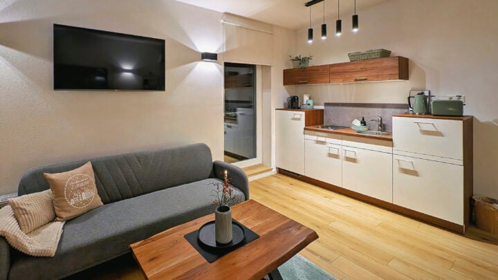 MainZauber Apartments früher MainTerrasse Miltenberg am Main - Erleben Sie den ultimativen Komfort in einer Ferienwohnung mit unserer luxuriösen Unterkunft. Dieser elegante Raum verfügt über einen gemütlichen, modernen Wohn- und Küchenbereich, perfekt für Ihren Kurzurlaub. Entspannen Sie auf dem stilvollen, grauen Sofa mit den weichen Kissen, während Sie Unterhaltung auf dem an der Wand montierten Fernseher genießen. Der Wohnbereich umfasst auch einen charmanten Couchtisch aus Holz, der ein anspruchsvolles und dennoch einladendes Ambiente schafft.
Unsere Luxuswohnung verfügt über eine kompakte Küche mit schönen Holzschränken, ideal zum Zubereiten von Mahlzeiten während Ihres Aufenthalts. Sanfte Beleuchtung sorgt überall für eine warme und einladende Atmosphäre und verbessert Ihr Entspannungserlebnis. Eine Tür führt bequem in ein weiteres Zimmer und bietet Ihnen zusätzlichen Platz zum Entspannen während Ihres Traumurlaubs in diesem exquisiten Rückzugsort. Entdecken Sie noch heute unvergleichliche Gemütlichkeit und Stil in unserer erstklassigen Ferienunterkunft!