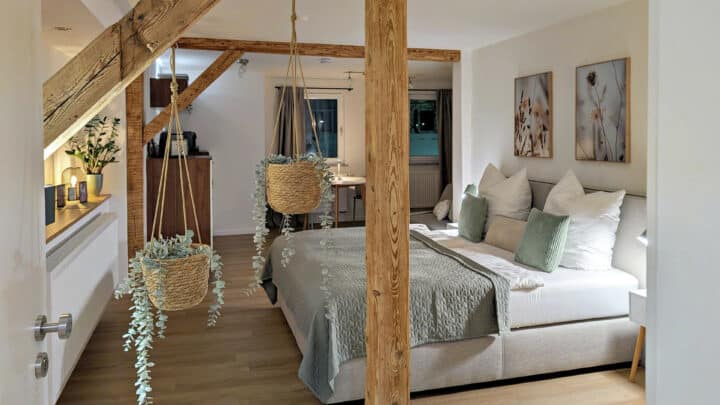 MainZauber Apartments früher MainTerrasse Miltenberg am Main - Erleben Sie die perfekte Mischung aus Komfort und Stil in unserer luxuriösen Ferienwohnung. Dieses gemütliche Schlafzimmer mit rustikalen Holzbalken und modernem Dekor bietet einen erholsamen Rückzugsort für Ihren Urlaub. Genießen Sie eine erholsame Nachtruhe auf dem großen Bett mit weichen grünen und weißen Kissen. Der Charme des Zimmers wird durch zwei hängende Pflanzenkörbe und geschmackvolle Wandkunst verstärkt, die eine einladende Atmosphäre schaffen. Warme Beleuchtung ergänzt den Raum, während eine praktische Küchenzeile im Hintergrund dafür sorgt, dass all Ihre Bedürfnisse während Ihres Aufenthalts erfüllt werden. Buchen Sie diese exquisite Unterkunft jetzt für ein unvergessliches Urlaubserlebnis.