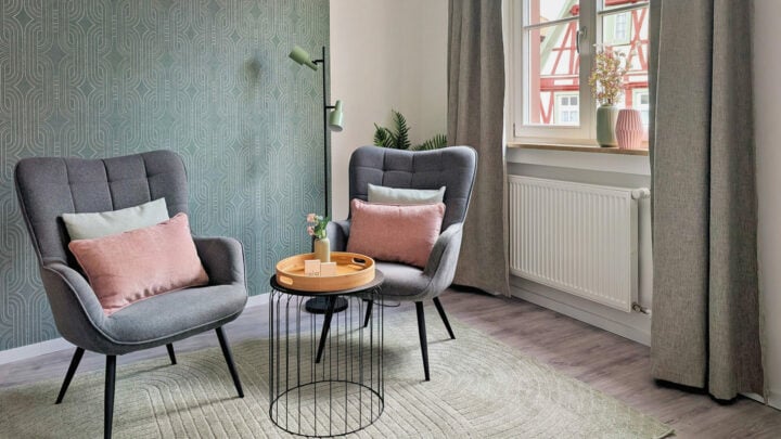 MainZauber Apartments früher MainTerrasse Miltenberg am Main - Gemütliches Luxus-Apartment: Moderne graue Sessel und stilvoller Tisch, große Fenster und grüne Tapete in zentraler Altstadtlage.