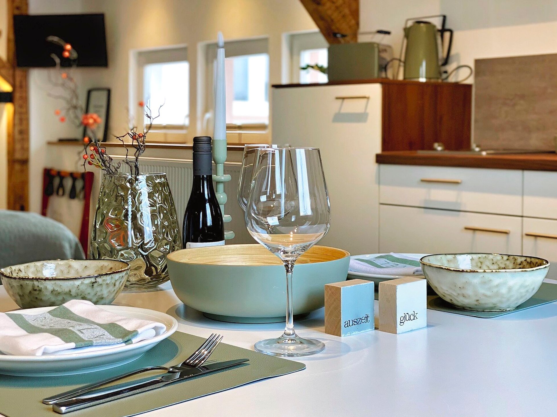 MainZauber Apartments Miltenberg am Main - Erleben Sie den ultimativen Komfort in dieser luxuriösen Ferienwohnung, in der ein gut gedeckter Esstisch die Mahlzeiten in eine elegante Angelegenheit verwandelt. Diese geräumige und gemütliche Wohnküche verfügt über elegante weiße Schränke und stilvolle Holzregale, die eine warme Atmosphäre schaffen, perfekt für Ihre Urlaubsunterkunftsbedürfnisse. Genießen Sie intime Abendessen mit grünen Schüsseln, einem Weinglas und einer Flasche Wein, ergänzt durch geschmackvolle Dekorationselemente. Ihre ideale Ferienwohnung erwartet Sie – buchen Sie jetzt, um sich während Ihres Urlaubs Entspannung und Eleganz zu gönnen.