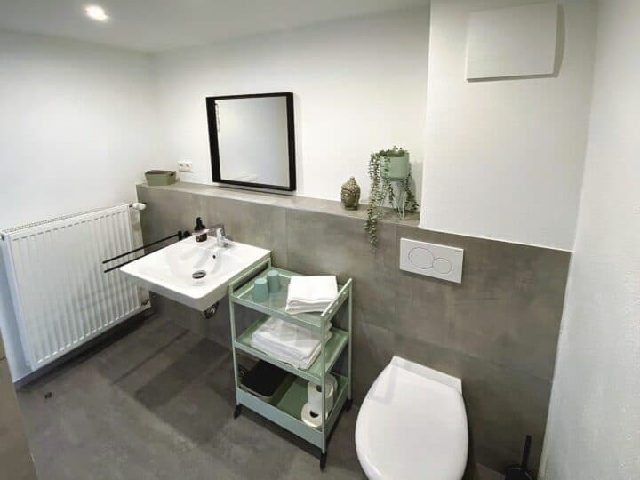 MainZauber Apartments früher MainTerrasse Miltenberg am Main - Entdecken Sie das ultimative Luxusapartment für Ihren Urlaub in der Nähe von Miltenberg. Dieses stilvolle und moderne Badezimmer, Teil Ihrer idealen Urlaubsunterkunft, besticht durch glatte weiße Wände und eine minimalistische Ästhetik. Genießen Sie den Komfort eines wandmontierten Waschbeckens mit Spiegel und einem Heizkörper, der für Gemütlichkeit sorgt. Ein eleganter Servierwagen ist sorgfältig mit flauschigen Handtüchern und hochwertigen Toilettenartikeln bestückt, während eine geschmackvolle Kunstpflanze einen erfrischenden Hauch von Grün in dieser exquisiten Main-Location verleiht. Sichern Sie sich jetzt Ihr Traum-Ferienapartment für einen unvergleichlichen Kurzurlaub!