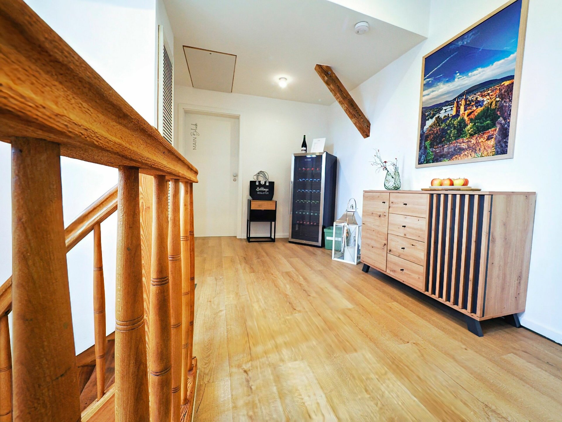 Treten Sie ein in die Ferienwohnung Ihrer Träume, wo Sie ein moderner und heller Flur mit einer eleganten Holztreppe und einem schicken Sideboard begrüßt. Dieser exquisite Raum, der sich perfekt für Ihren Luxusapartmenturlaub eignet, verfügt über einen praktischen Weinkühlschrank, der Ihren Aufenthalt noch schöner macht. Die makellos weiße Wand ziert ein atemberaubendes Landschaftskunstwerk, begleitet von einer stilvollen Anordnung frischer Blumen in einer Vase und einer Schale mit Äpfeln auf dem Sideboard – jedes Detail ist für eine unvergessliche Urlaubsunterkunft konzipiert. - MainZauber Apartments Miltenberg am Main