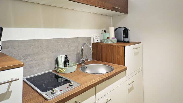 MainZauber Apartments früher MainTerrasse Miltenberg am Main - Erleben Sie den Komfort und die Bequemlichkeit unserer luxuriösen Ferienwohnung, perfekt für Ihren nächsten Urlaub. Unsere voll ausgestattete Kompaktküche verfügt über elegante Holzarbeitsplatten und einen modernen Elektroherd, sodass Sie während Ihres Aufenthalts alles haben, was Sie brauchen, um köstliche Mahlzeiten zuzubereiten. Neben der schicken runden Spüle steht eine praktische Kaffeemaschine für Ihren Morgenkaffee. Durchdachte Details wie grüne Tabletts, auf denen Utensilien und Spülmittel ordentlich aufbewahrt werden, tragen zum organisierten Charme dieser ansprechenden Unterkunft bei. Ein hübsches Schild neben dem Wasserhahn verleiht dekoratives Flair und unterstreicht die einladende Atmosphäre dieser exquisiten Ferienwohnung.