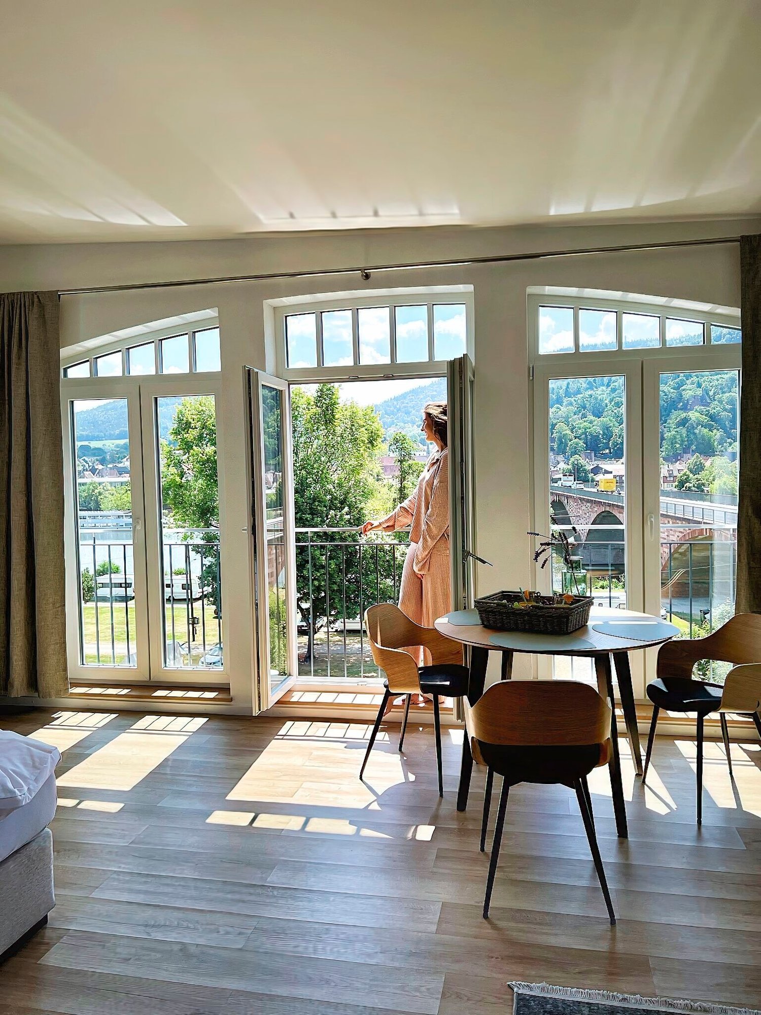 Stellen Sie sich vor, Sie stehen auf dem Balkon einer luxuriösen Ferienwohnung und genießen die atemberaubende Aussicht auf einen ruhigen Fluss und sanfte Hügel. Diese stilvolle Unterkunft verfügt über große Fenster, die einen atemberaubenden Panoramablick bieten und natürliches Licht in das gemütliche Interieur bringen. Genießen Sie köstliche Mahlzeiten an Ihrem runden Esstisch mit vier bequemen Stühlen, alles vor eleganten Hartholzböden. Genießen Sie die perfekte Mischung aus Komfort und Ruhe in Ihrem Traumurlaubsort. - MainZauber Apartments Miltenberg am Main