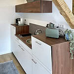 Erleben Sie die perfekte Mischung aus Stil und Komfort in dieser luxuriösen Ferienwohnung. Diese atemberaubende Unterkunft verfügt über eine kompakte, aber gut gestaltete Küche mit eleganten weißen und hölzernen Schränken. Genießen Sie die Zweckmäßigkeit einer kleinen Spüle, eines elektrischen Kochfelds und eines charmanten grünen Toasters neben Ihrer Morgenkaffeemaschine. Ein Hauch von Natur ziert die Arbeitsplatte mit einer schönen Pflanze und verstärkt die ruhige Atmosphäre. Der gemütliche Holzboden ist teilweise mit einem eleganten Teppich geschmückt, während rustikale Holzbalken diesem außergewöhnlichen Rückzugsort Charakter verleihen. Perfekt für Reisende, die Komfort in ihrer Ferienwohnung suchen! - MainZauber Apartments Miltenberg am Main