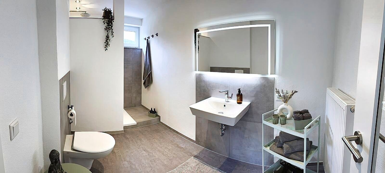 Erleben Sie einen luxuriösen Aufenthalt im modernen Badezimmer dieser Ferienwohnung, das Ihren Aufenthalt noch angenehmer macht. Schwelgen Sie in dem elegant eingerichteten Raum mit glatten weißen Wänden und schicken grauen Fliesenböden. Das Badezimmer ist mit einem stilvollen Waschbecken und einem großen Spiegel sowie einer modernen ebenerdigen Dusche und Toilette ausgestattet, sodass all Ihre Komfortbedürfnisse erfüllt werden. Genießen Sie den Komfort ordentlich angeordneter Toilettenartikel auf einem kleinen Tisch und flauschiger Handtücher, die in Reichweite hängen. Die helle Beleuchtung erhellt diese gehobene Umgebung wunderschön und verleiht Ihrem Luxusapartment für den Urlaub einen zusätzlichen Hauch von Eleganz. - MainZauber Apartments Miltenberg am Main