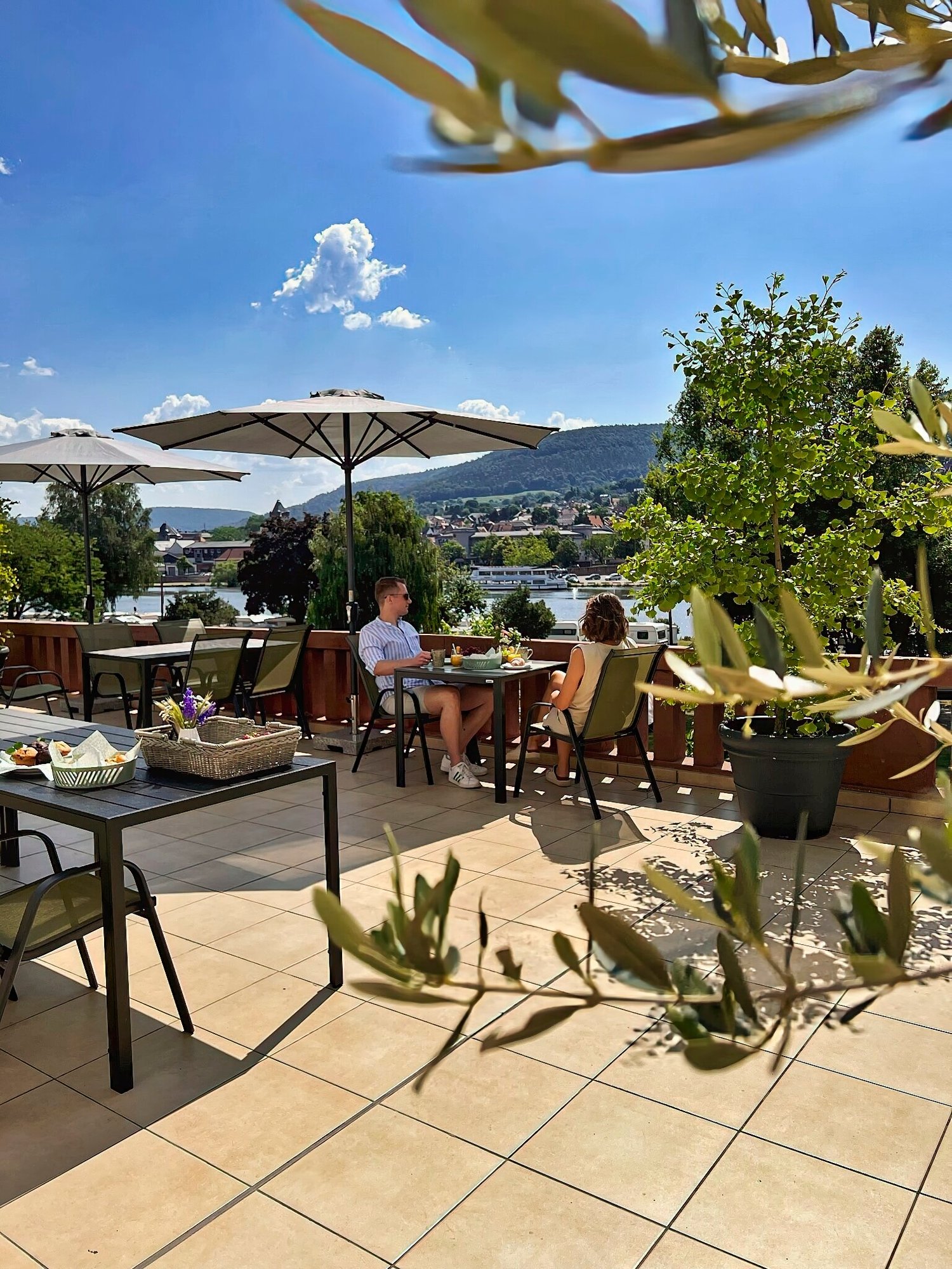 MainZauber Apartments Miltenberg am Main - Stellen Sie sich vor, Sie entspannen sich in einer Ferienwohnung und genießen einen entspannten Morgen auf der sonnigen Außenterrasse. Diese luxuriöse Unterkunft verfügt über Sonnenschirme und Topfpflanzen und schafft so eine einladende Atmosphäre für Ihren Urlaub. Die Aussicht bietet einen atemberaubenden Blick auf einen malerischen Fluss und üppige Hügel unter einem klaren blauen Himmel mit flauschigen Wolken. In der Nähe sind charmante Tische elegant mit Tabletts und Blumen gedeckt und unterstreichen die ruhige Atmosphäre Ihres perfekten Urlaubsdomizils.