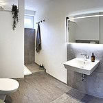 Erleben Sie einen luxuriösen Aufenthalt im modernen Badezimmer dieser Ferienwohnung, das Ihren Aufenthalt noch angenehmer macht. Schwelgen Sie in dem elegant eingerichteten Raum mit glatten weißen Wänden und schicken grauen Fliesenböden. Das Badezimmer ist mit einem stilvollen Waschbecken und einem großen Spiegel sowie einer modernen ebenerdigen Dusche und Toilette ausgestattet, sodass all Ihre Komfortbedürfnisse erfüllt werden. Genießen Sie den Komfort ordentlich angeordneter Toilettenartikel auf einem kleinen Tisch und flauschiger Handtücher, die in Reichweite hängen. Die helle Beleuchtung erhellt diese gehobene Umgebung wunderschön und verleiht Ihrem Luxusapartment für den Urlaub einen zusätzlichen Hauch von Eleganz. - MainZauber Apartments Miltenberg am Main