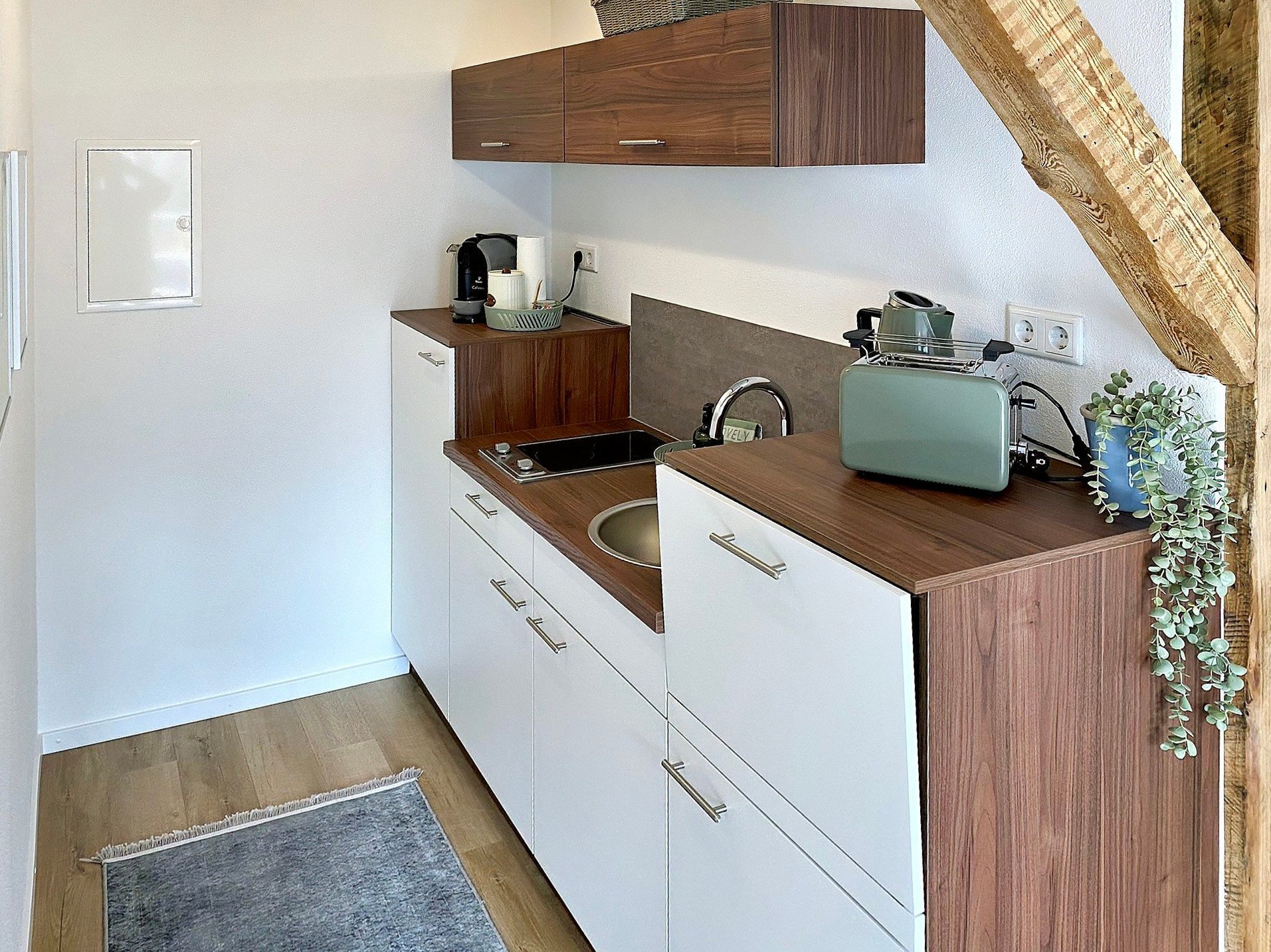 Erleben Sie die perfekte Mischung aus Stil und Komfort in dieser luxuriösen Ferienwohnung. Diese atemberaubende Unterkunft verfügt über eine kompakte, aber gut gestaltete Küche mit eleganten weißen und hölzernen Schränken. Genießen Sie die Zweckmäßigkeit einer kleinen Spüle, eines elektrischen Kochfelds und eines charmanten grünen Toasters neben Ihrer Morgenkaffeemaschine. Ein Hauch von Natur ziert die Arbeitsplatte mit einer schönen Pflanze und verstärkt die ruhige Atmosphäre. Der gemütliche Holzboden ist teilweise mit einem eleganten Teppich geschmückt, während rustikale Holzbalken diesem außergewöhnlichen Rückzugsort Charakter verleihen. Perfekt für Reisende, die Komfort in ihrer Ferienwohnung suchen! - MainZauber Apartments Miltenberg am Main
