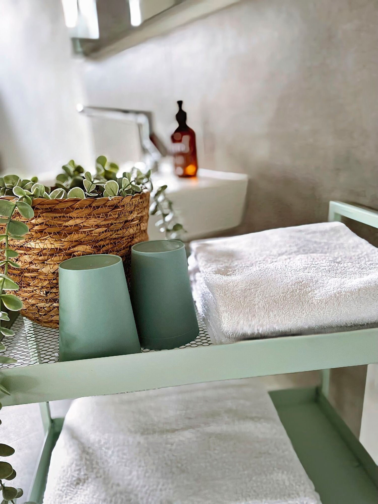 Entdecken Sie in unserer luxuriösen Ferienwohnung einen ruhigen und Spa-ähnlichen Rückzugsort, perfekt für Ihren nächsten Kurzurlaub. Stellen Sie sich vor, Sie entspannen sich in einem elegant gestalteten Badezimmer mit einem stilvollen grünen Metallwagen, der mit tadellos gefalteten weißen Handtüchern bestückt ist. Für zusätzlichen Komfort finden Sie zwei bezaubernde grüne Tassen und einen geflochtenen Korb, der mit erfrischendem Grün geschmückt ist. Beachten Sie im sanft verschwommenen Hintergrund das glatte Waschbecken und die elegante braune Flasche, die das ruhige Ambiente dieser exquisiten Unterkunft unterstreichen. Gönnen Sie sich das ultimative Entspannungserlebnis, wenn Sie diese außergewöhnliche Ferienwohnung für Ihren Aufenthalt buchen. - MainZauber Apartments Miltenberg am Main